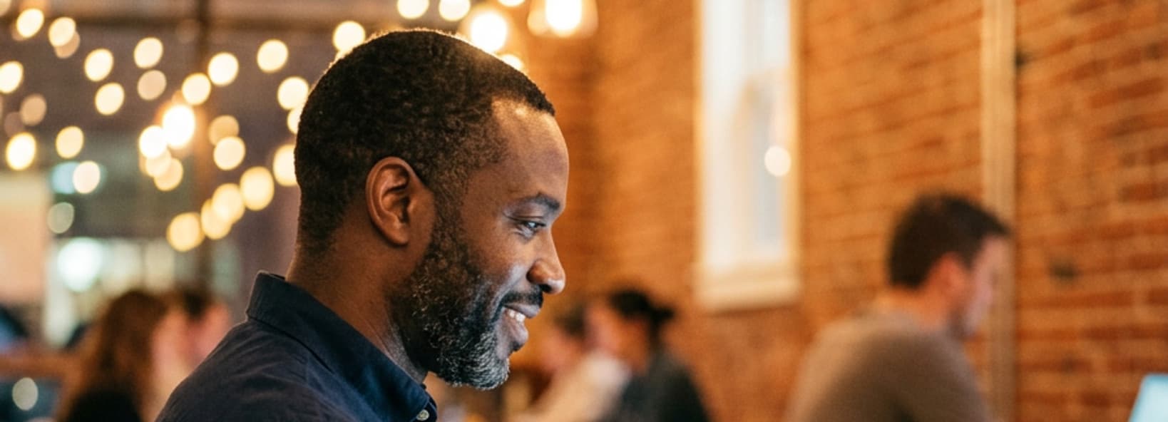 Man with a short beard in a navy shirt working on a laptop in a warm, brick-walled coworking space