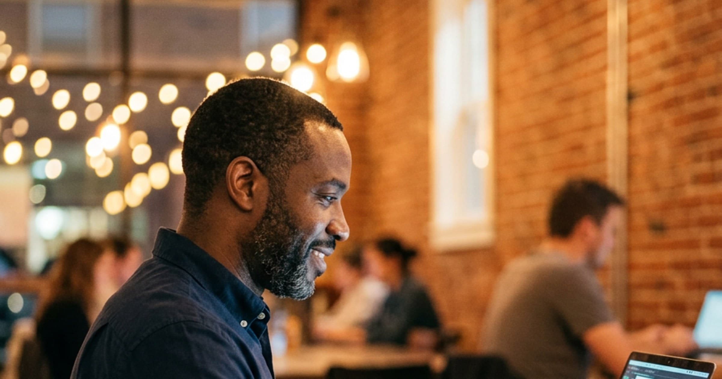 Man with a short beard in a navy shirt working on a laptop in a warm, brick-walled coworking space