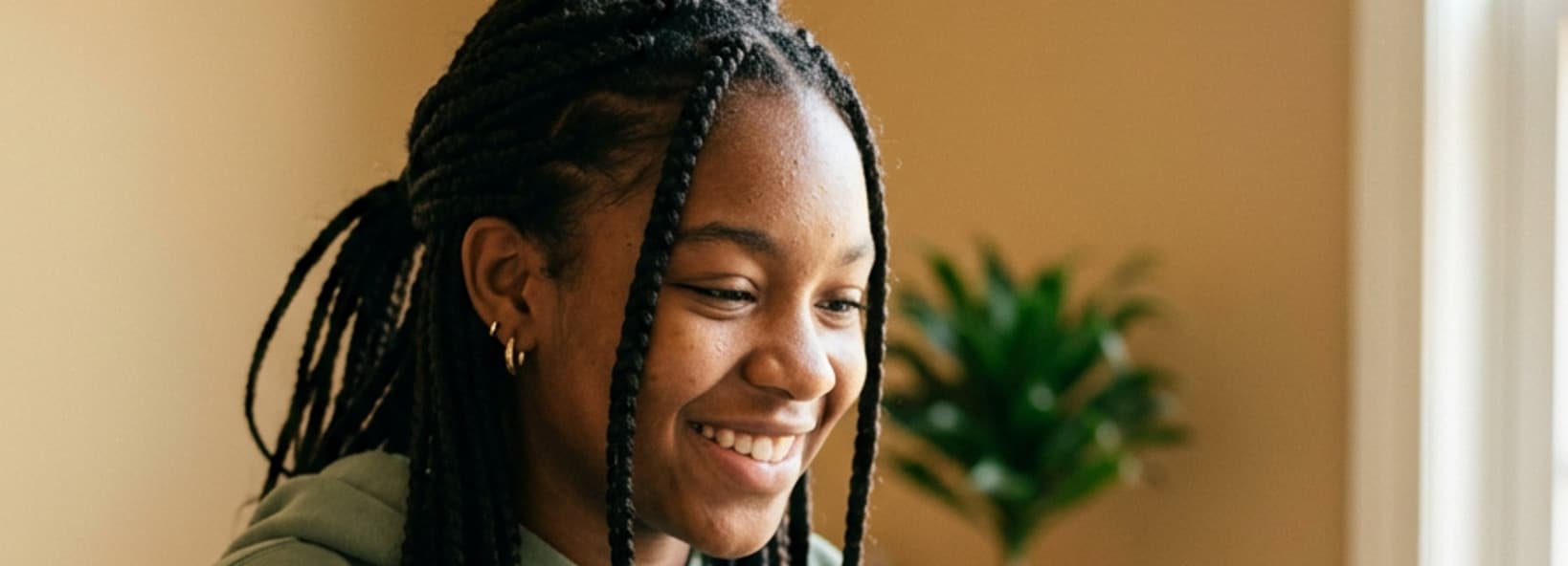 Smiling teenage girl with braids in a green hoodie, in a warm room with a plant