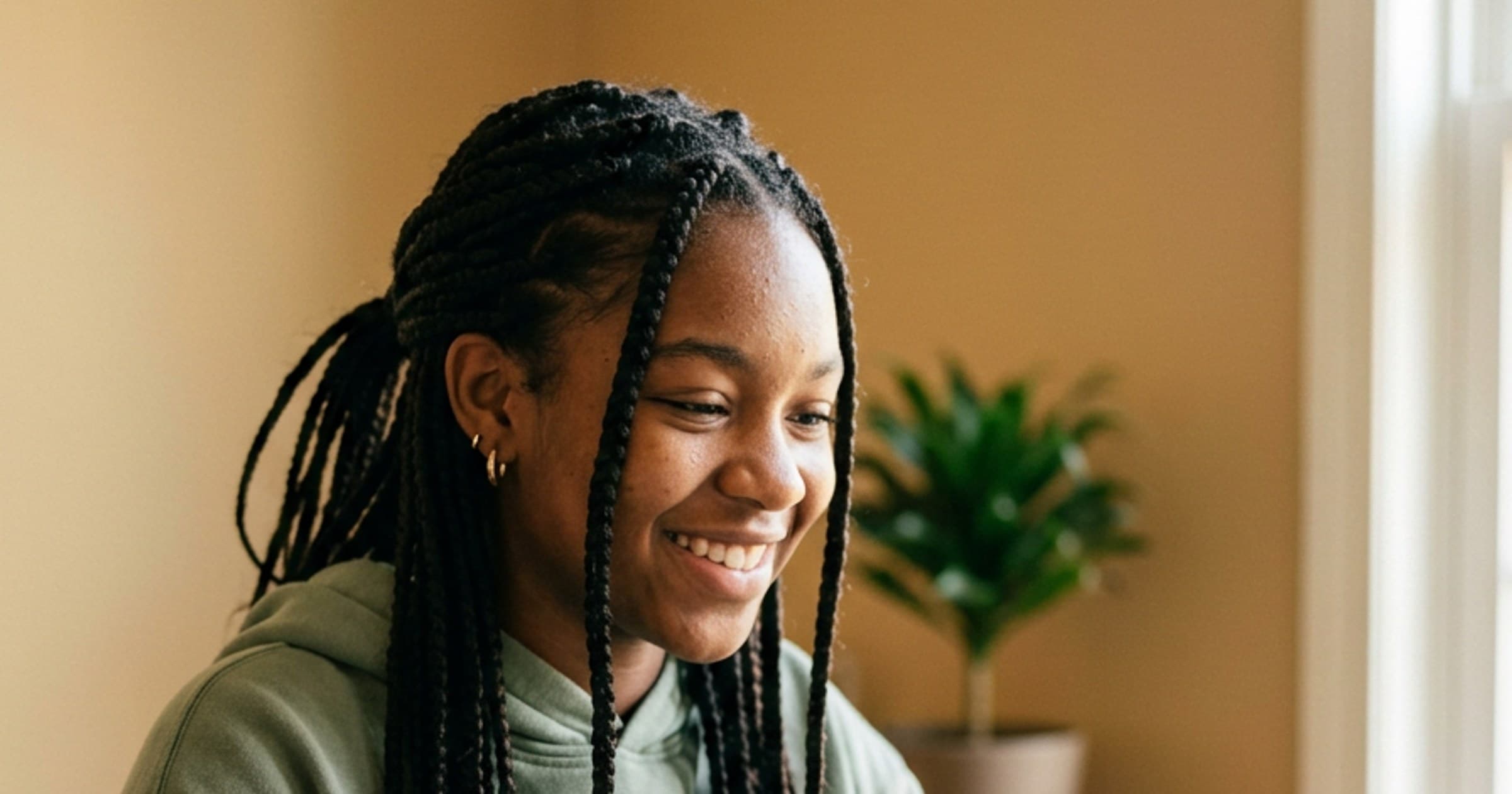 Smiling teenage girl with braids in a green hoodie, in a warm room with a plant